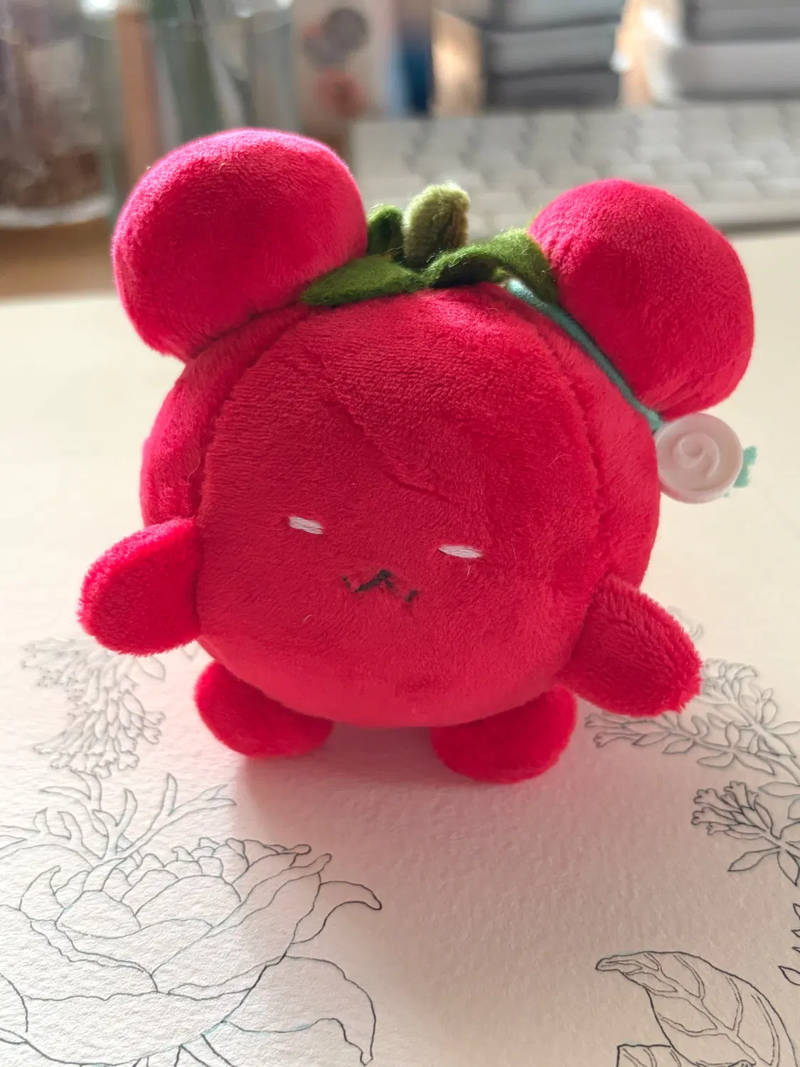 丸い体に丸い耳がふたつと小さい手足がついた赤い生き物「トマチュ」のぬいぐるみ写真。頭には緑のヘタがついている